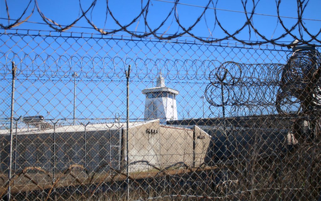 A grey concrete building with guard tower, seen through razor wire fencing.