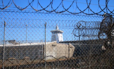A grey concrete building with guard tower, seen through razor wire fencing.