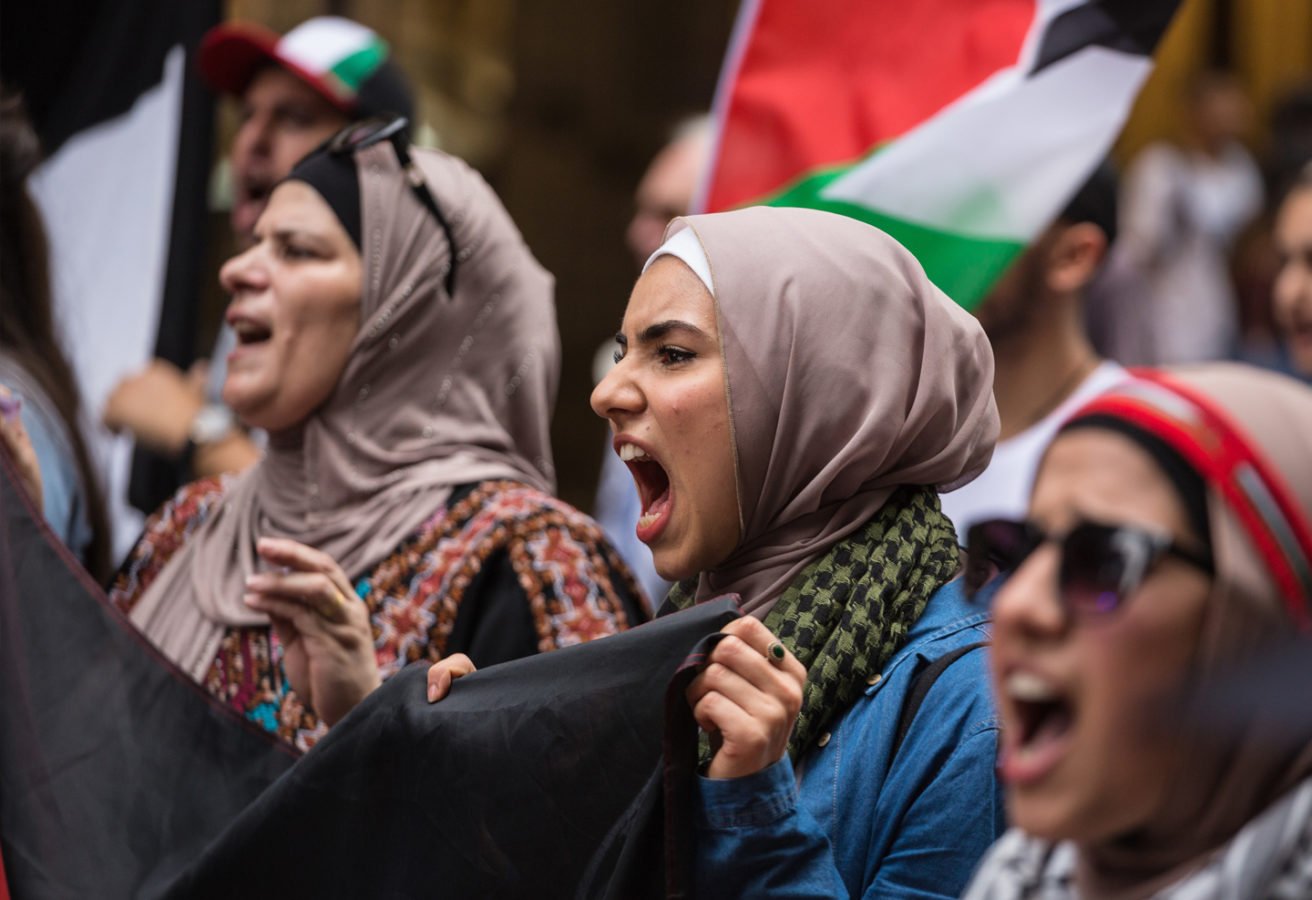 Women in Muslim and traditional Palestinian costume holding a banner and chanting slogans.