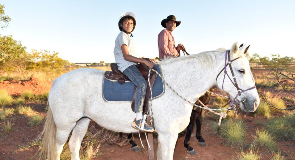 A boy rides a horse with co-founder David Sammon as part of Mona’s Cultural Horsemanship program on Flora Downs Station, near Mount Isa, June 2016. © Wayne Quilliam