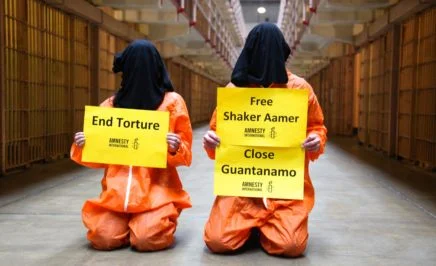 two people in Guantanamo Bay style orange jumpsuits - one holding a sign that says End Torture; the other holding a sign which says that says Free Shaker Aamer and Close Guantanamo.