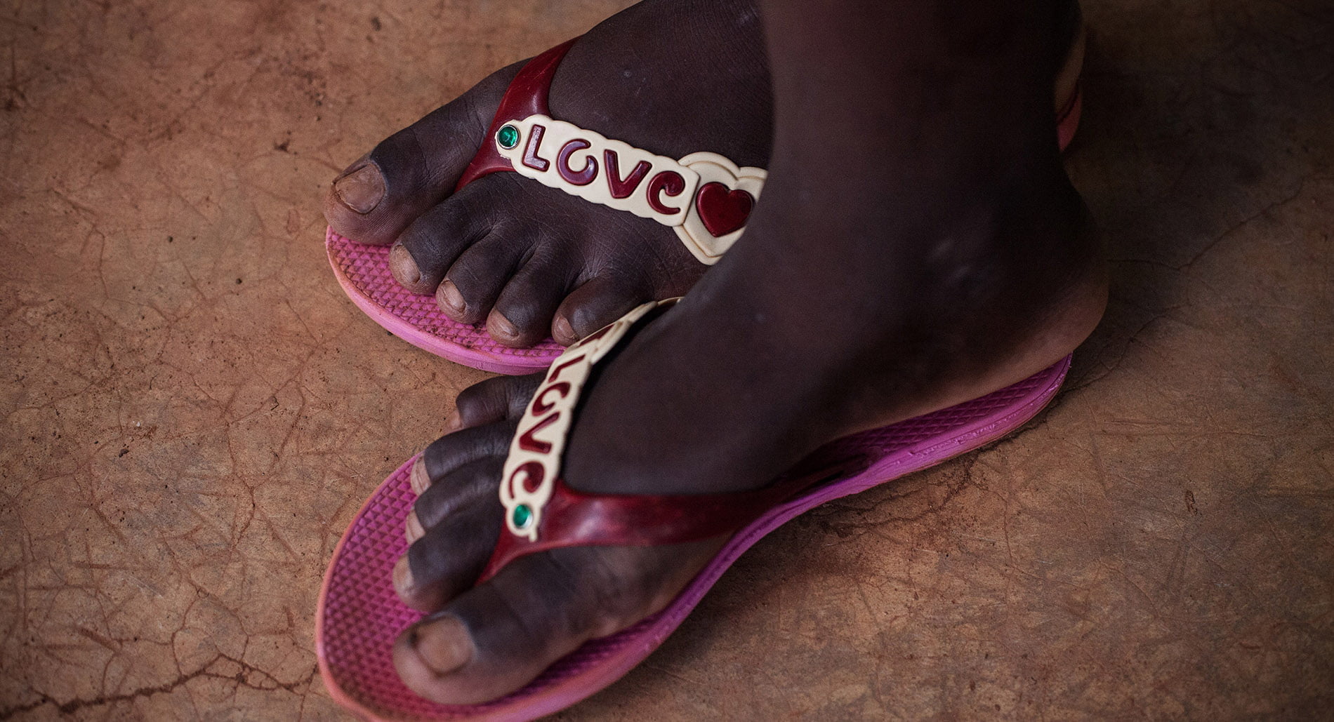two feet with bright pink plastic sandals with 'love' written on them, standing on red soil