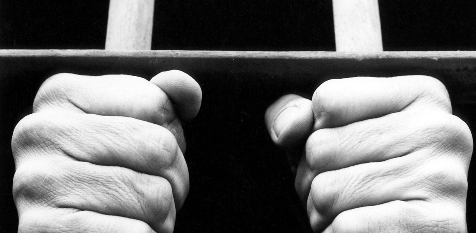Black and white photograph showing hands of an anonymous prisoner gripping the bars of a prison cell.