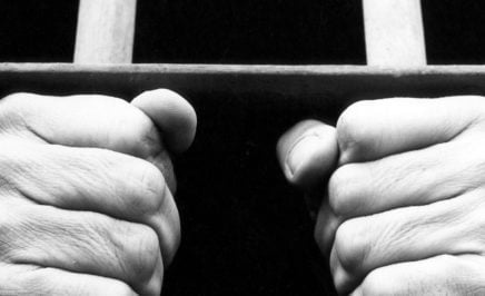 Black and white photograph showing hands of an anonymous prisoner gripping the bars of a prison cell.