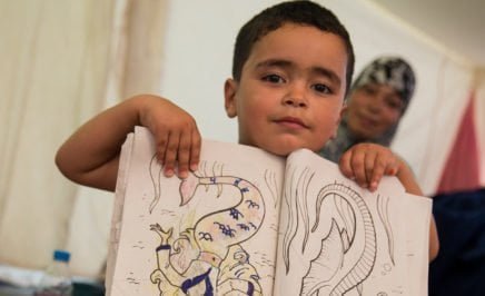 A young refugee staying in a camp in Greece holds his colouring-in book up to show the camera