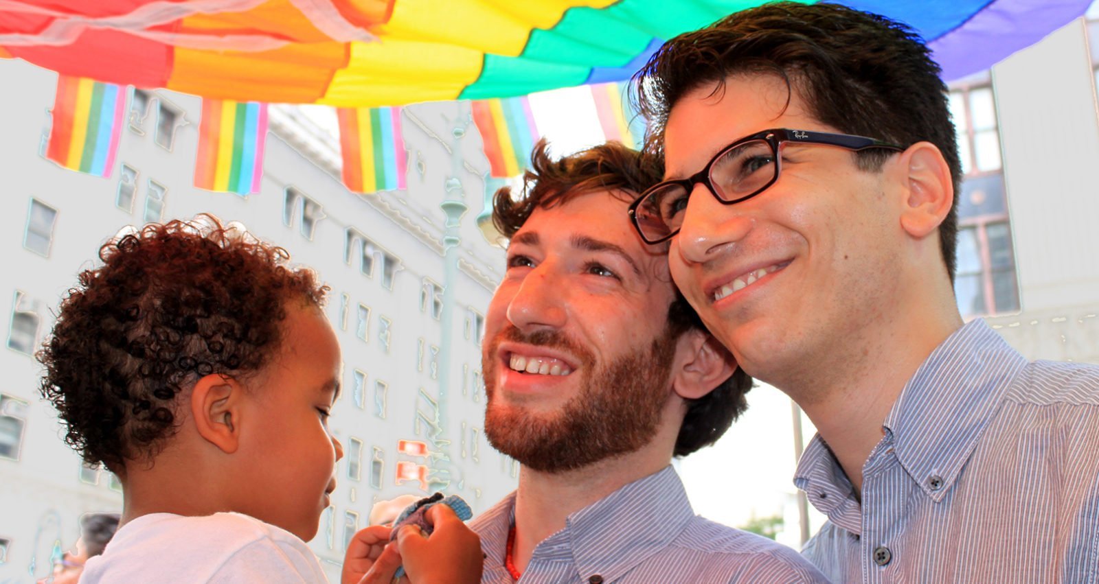 Two men standing together, smiling and holding a young child under a rainbow flag.