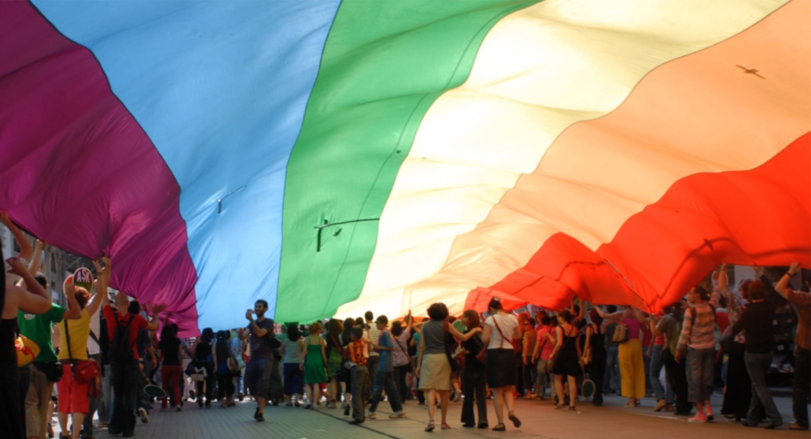 A large group of people standing under a rainbow flag.