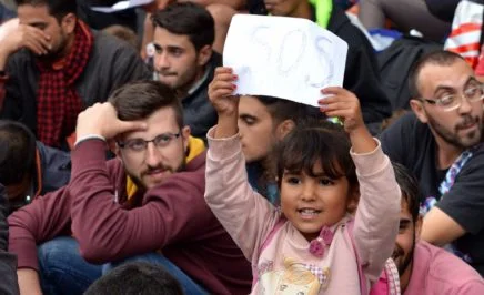 child holds up a sign with SOS written on it, while standing in a crowd of people