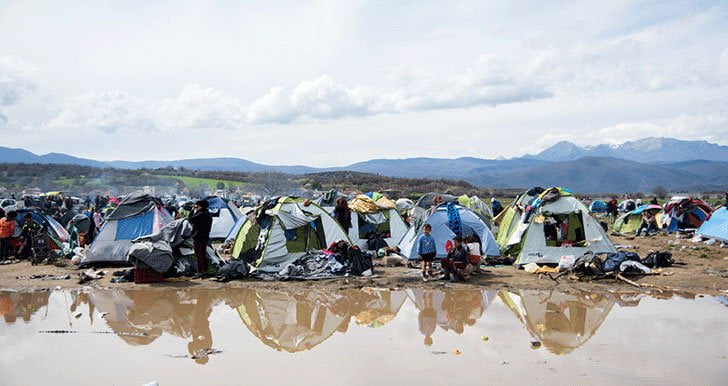 A refugee camp with a crowd of tents near a puddle of muddy water