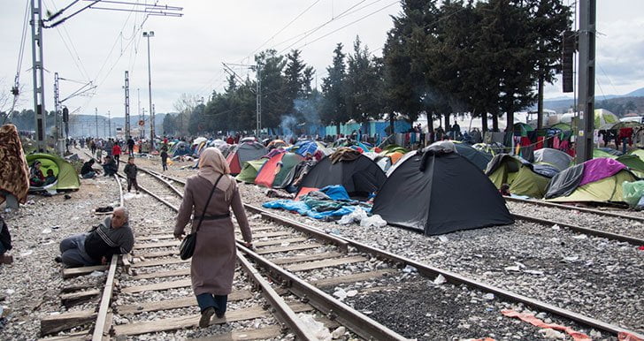 A woman walking along train tracks away from the camera. To the right of the tracks are refugee camp tents.