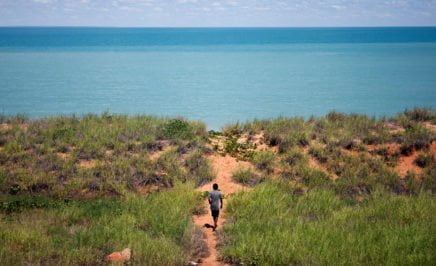 A young Indigenous man walking through the bush toward the ocean.
