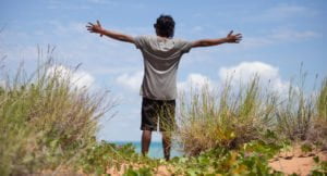 A young Indigenous man with his arms outstretched toward the ocean
