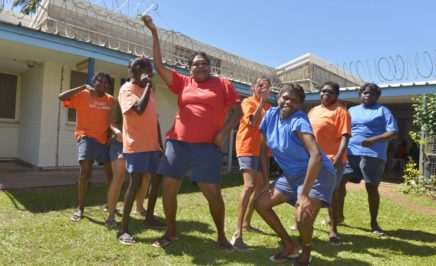 Still from documentary prison songs in which several indigenous women are dancing in front of a white building with barbed wire along the gutters