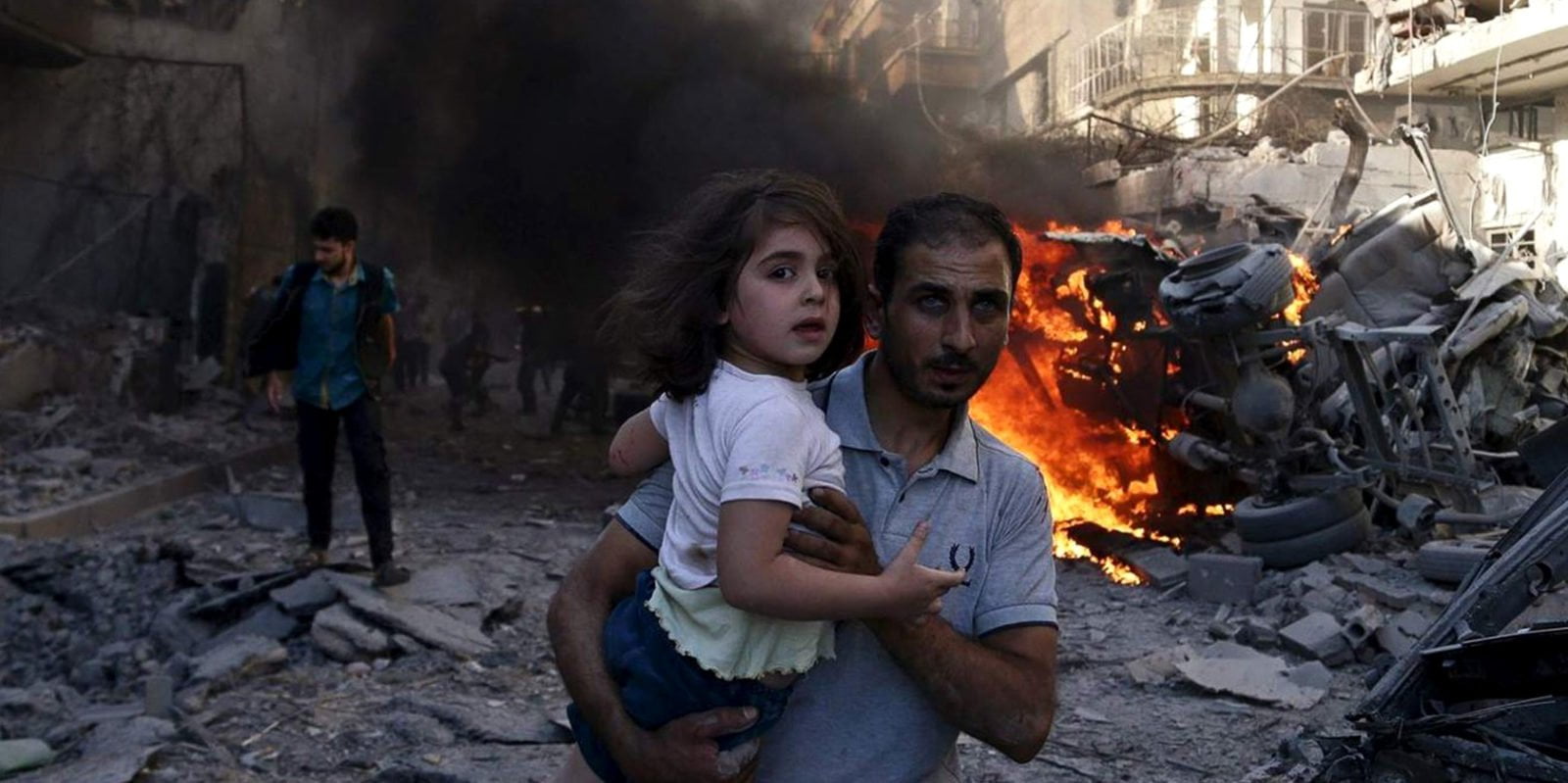 A man carries a young girl through rubble in Syria.