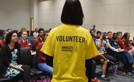 Person in a yellow amnesty volunteer tshirt stands in front of a crowd of seated people