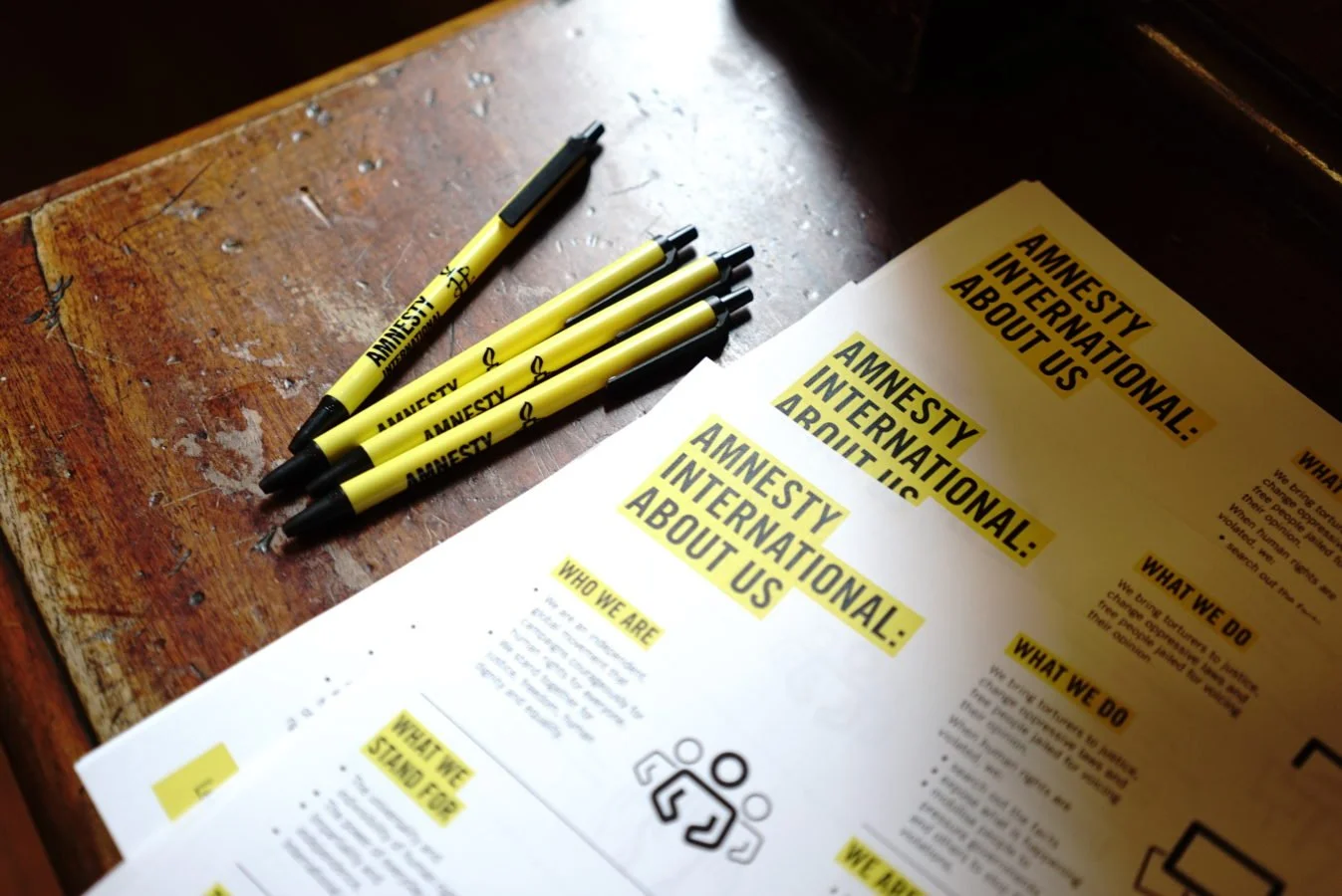 Flyer titled 'Amnesty International: About Us' on a desk with Amnesty branded pens
