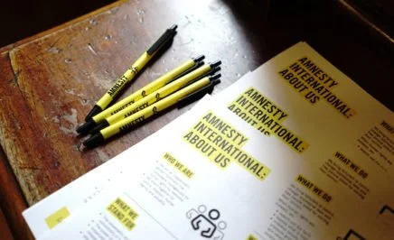 Flyer titled 'Amnesty International: About Us' on a desk with Amnesty branded pens
