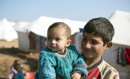 A smiling boy holds a baby, with white tents behind him