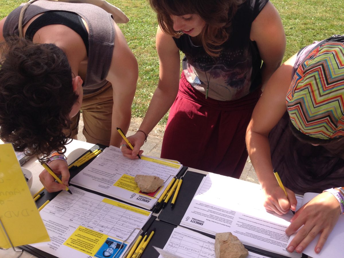 People lean over a table holding yellow Amnesty pens and sign a petition. They are in an outside setting, and grass is visible behind them.