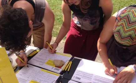 People lean over a table holding yellow Amnesty pens and sign a petition. They are in an outside setting, and grass is visible behind them.