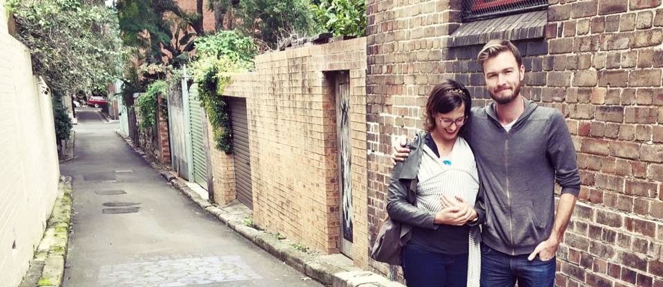An image of a woman and a man walking down a side street. The man has his arm around the woman and the woman is wearing a newborn baby in a sling.