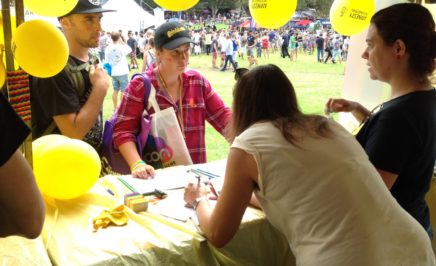 Volunteers stand behind an Amnesty International Stall at Mardi Gras - the table is decorated with yellow table-cloth, and yellow balloons hang above. The volunteers are pictured speaking to interested people about a campaign.
