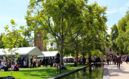 Students surround the Melbourne University Campus - the grassy south lawn and a small body of water can be seen on a sunny day. There are white stalls that are filled with information for new students to explore.