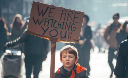 Young boy holding sign which says 'we are watching you'