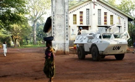 A woman passes in the foreground, a UN armoured vehicle stands in front of a church
