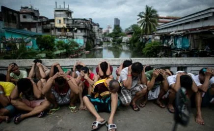 Drug suspects in the Philippines sit on the ground with their heads down, a canal and city skyline in the background
