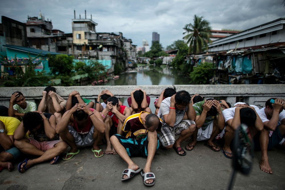 Drug suspects in the Philippines sit on the ground with their heads down, a canal and city skyline in the background
