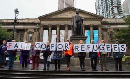 A group of people stand in front of the Victorian State Library, holding cards that spell out 'Fair go for refugees'