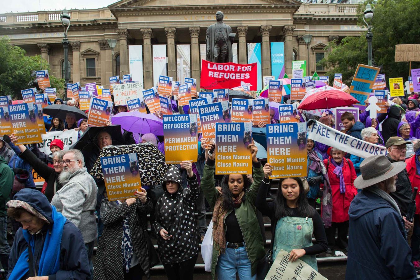 Melbourne protestors stand outside the State Library of Victoria holding signs that read 'Bring them here'. It is a rainy day, so people wear raincoats and hold umbrellas.