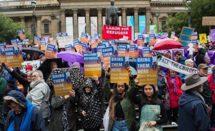 Melbourne protestors stand outside the State Library of Victoria holding signs that read 'Bring them here'. It is a rainy day, so people wear raincoats and hold umbrellas.
