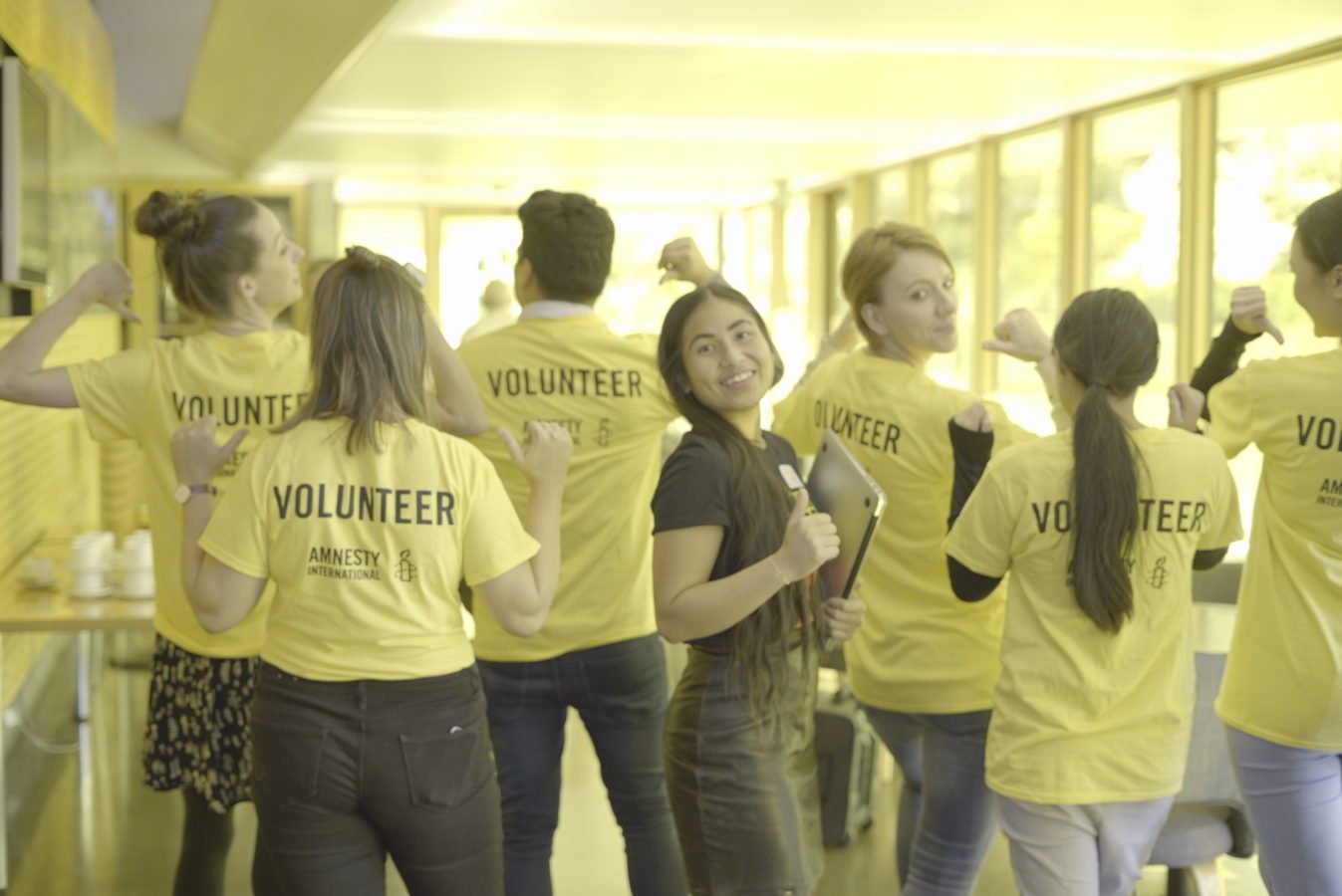 Volunteers stand with their backs to the camera, showing the backof their shirts that say 'volunteer'. They hold up their arms in a muscle flexing pose.