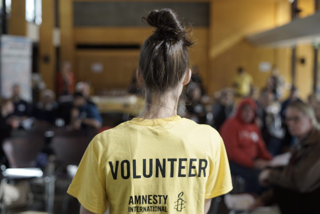 A shot of a volunteer addressing a crowd/ The shot is from behind. She is wearing a yellow shirt that says volunteer.