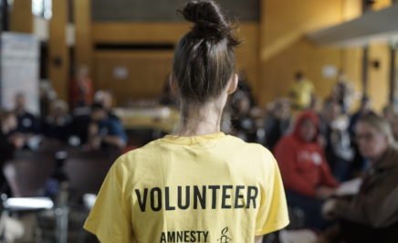 A shot of a volunteer addressing a crowd/ The shot is from behind. She is wearing a yellow shirt that says volunteer.