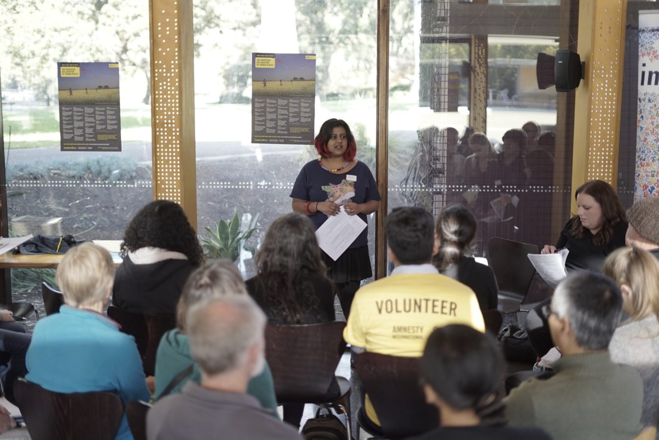 A woman stands in front of a seated audience. One audience member is weareing a yellow Amnesty shirt that reads 'volunteer'