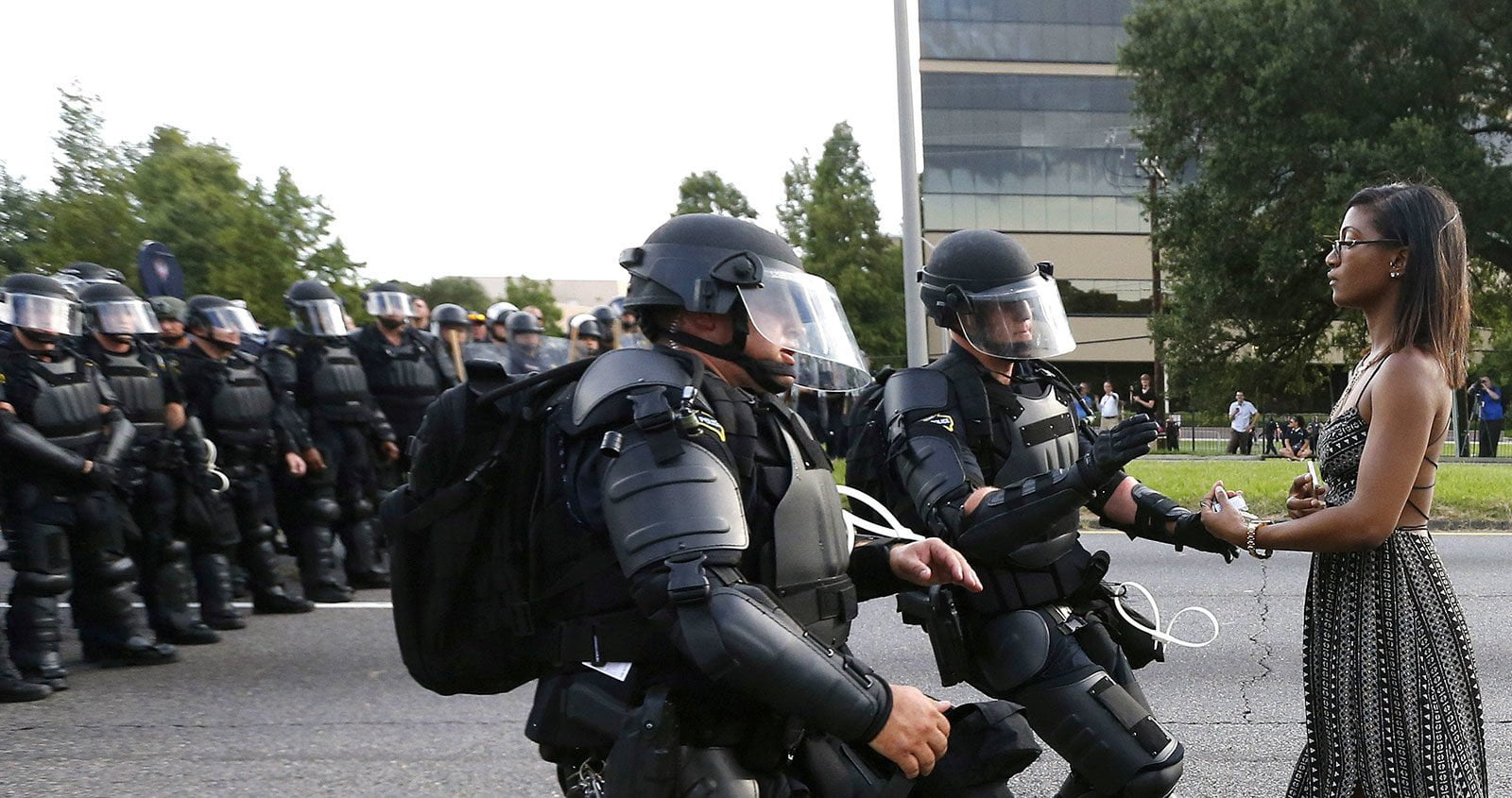A black female demonstrator stands still in the street as US police officers in riot gear rush toward her. Behind them is a line of police officers also in riot gear.