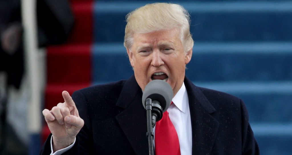 A photograph of President Donald Trump at his inauguration in January 2017. The image shows President Trump standing in front of a microphone in mid-sentence and pointing the index finger on his right hand towards the crowd (out of picture). Trump is wearing a dark coat, white shirt and red tie.