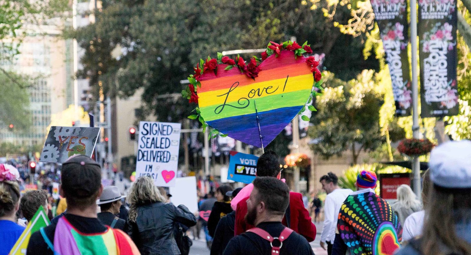 A photo of a crowd of people gather for a rally marriage equality rally in Sydney. There are numerous people holding rainbow flags and carrying signs that say 'Love!' and 'Vote Yes!'