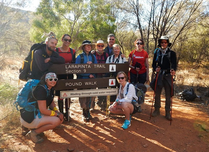 A photograph of the participants of the 2017 Inspired Adventures/Amnesty International Larapinta Trail trek. The image shows a group of men and women standing in the middle of the trail, surrounded by red earth and trees and standing next to a sign that says 'Larapinta Trail, Section 9, Pound Walk, Wading through cold water required.'