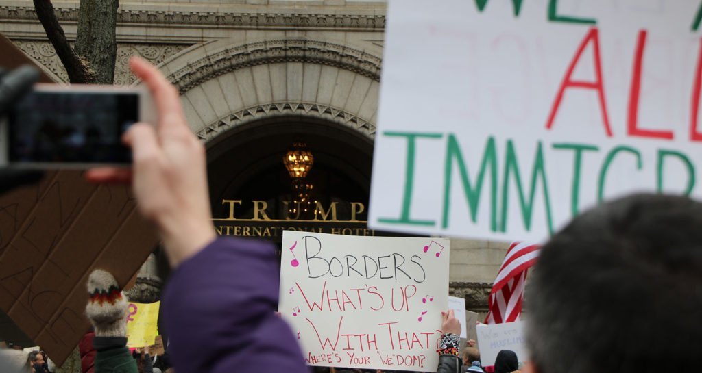 A photograph showing protesters outside Trump International Hotel in Washington DC. The image shows the backs of peoples' heads and arms in the air holding signs. The sign in shot says 'Borders: What's up with that? Where's your "WE"DOM'. The hotel signage can be in the foreground.