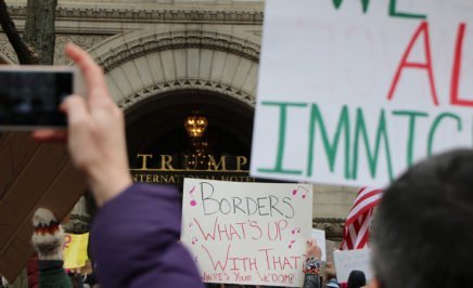 A photograph showing protesters outside Trump International Hotel in Washington DC. The image shows the backs of peoples' heads and arms in the air holding signs. The sign in shot says 'Borders: What's up with that? Where's your 
