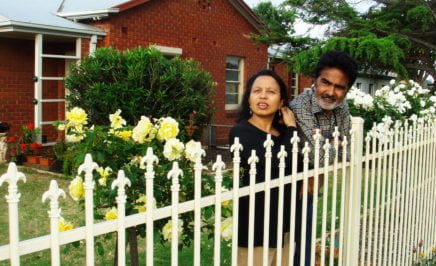 A photograph of human rights activist and former refugee from Bhutan Ratan Gazmere and his wife Gauri. The image shows the pair standing in their front garden standing near their white fence and looking into the distance. A red brick house is in the background.