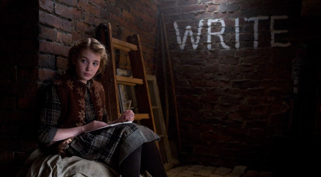 A still from the movie version of The Book Thief. The still shows actress Sophie Nélisse as Liesel Meminger, sitting in a basement reading. The word 'WRITE' is written in capitals on the wall behind her.