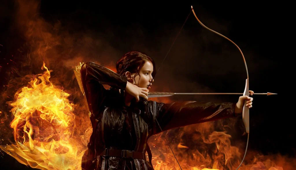 A still from the movie 'The Hunger Games'. The image shows Jennifer Lawrence as Katniss Everdeen, firing an arrow into the distance, flames in the background.