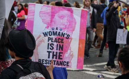 A photograph of a protester in a crowd of people at the women's march on Washington in January 2017. She is holding a sign featuring Carrie Fisher as Princess Leia from Star Wars with the caption 'A women's place is in the resistance'.