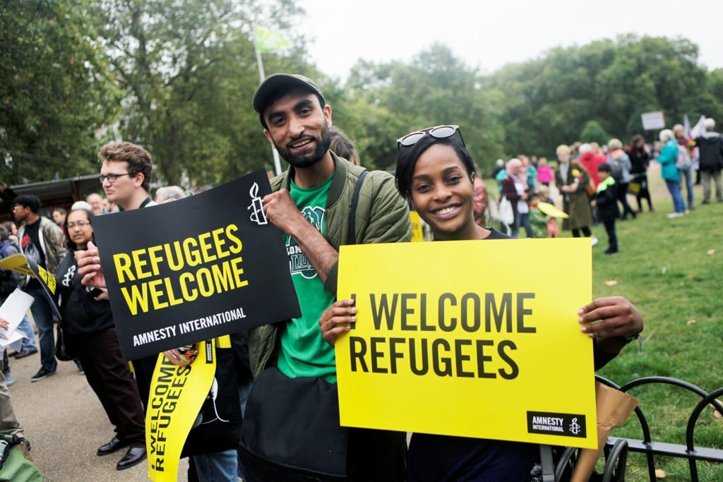 A man and woman at a rally in London to welcome refugees. They smile into the camera holding signs bearing the Amnesty International logo that say 'Refugees welcome'.