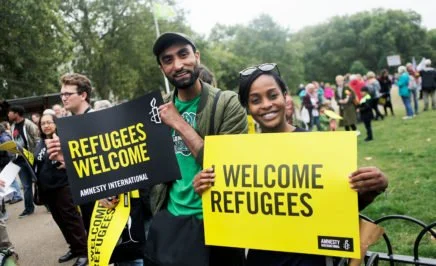 A man and woman at a rally in London to welcome refugees. They smile into the camera holding signs bearing the Amnesty International logo that say 'Refugees welcome'.